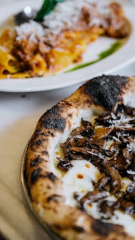 Close-up of a burnt crust pizza topped with mushrooms alongside a delicious pasta dish.