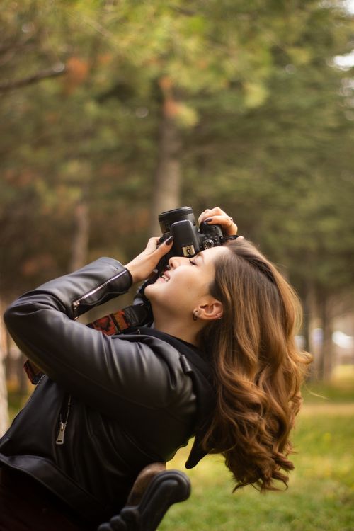 Free A young woman in a leather jacket taking pictures in a serene park setting, smiling and focused on her camera. Stock Photo