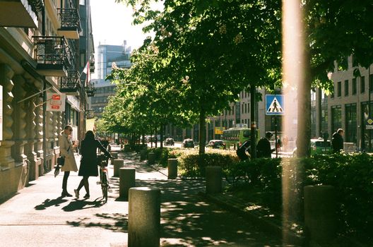 People walking on a sunlit Oslo sidewalk with trees and city buildings.