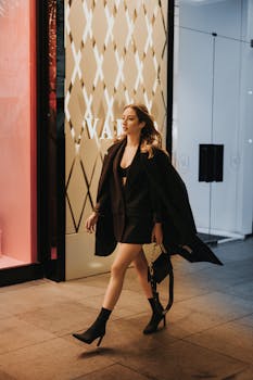 Fashionable woman in black attire confidently walking past a city storefront at night.