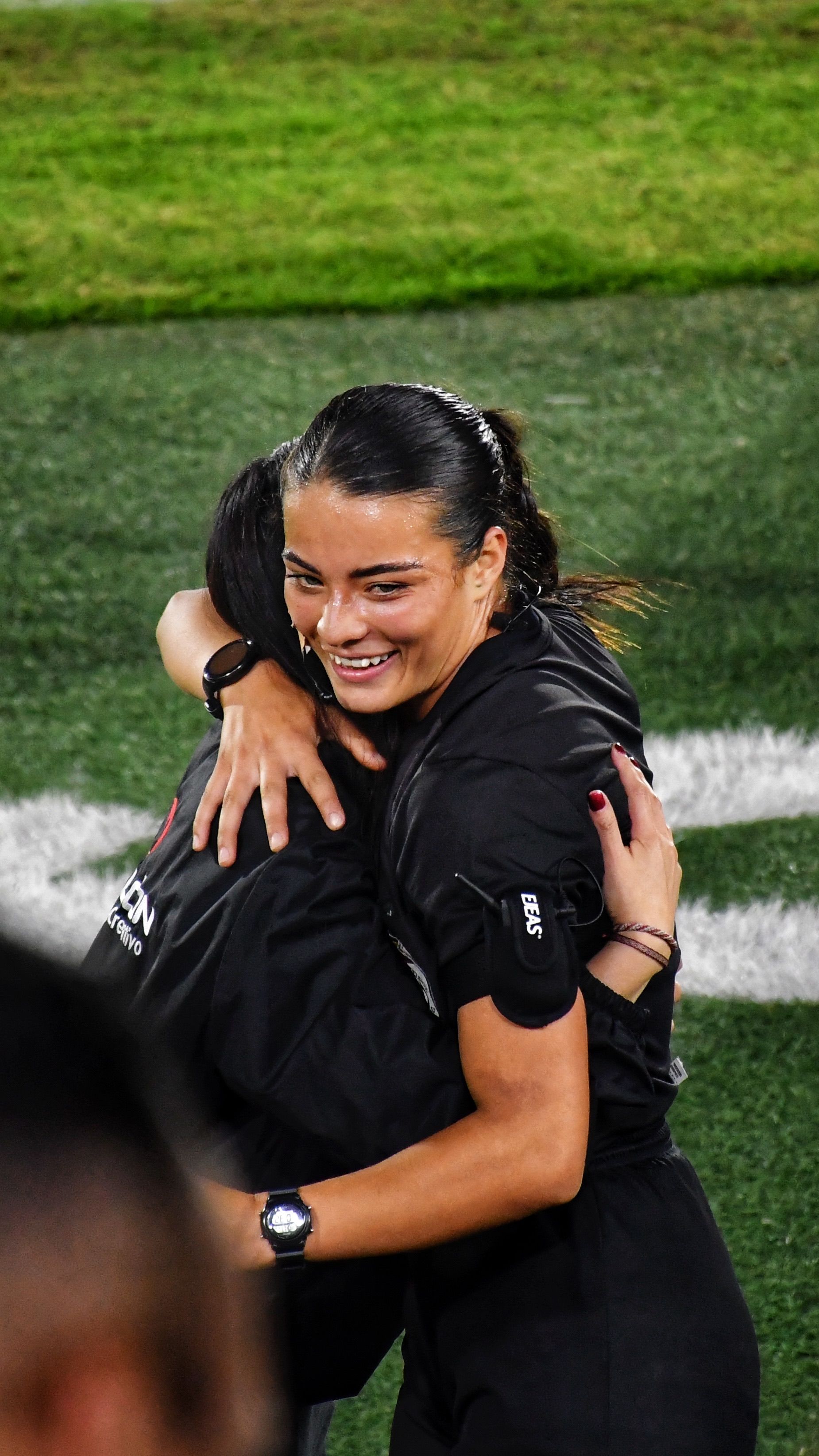 A female sports official joyfully hugs a colleague on a lush green field.