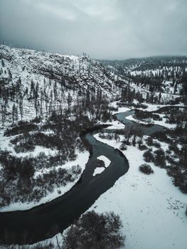 Aerial view of a black river winding through a snowy, mountainous winter landscape.