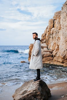 A stylish man poses in a coat on a rocky beach, blending fashion with nature's beauty.
