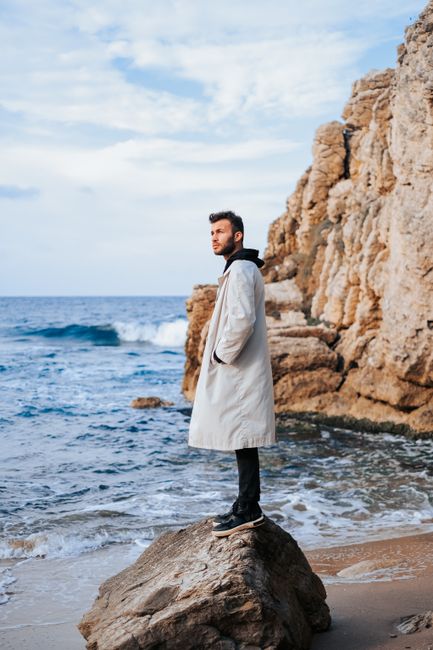 A stylish man poses in a coat on a rocky beach, blending fashion with nature's beauty.