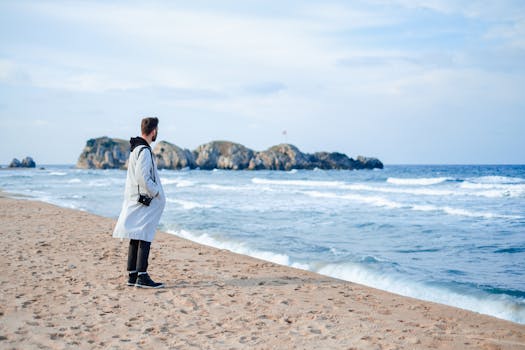A man in a jacket stands on a sandy beach, overlooking the ocean with rocky islands in the distance.