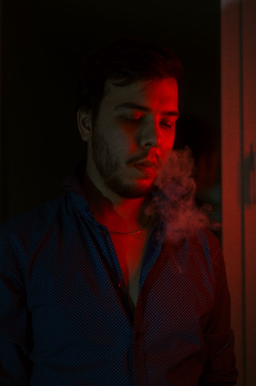 A man exhaling smoke, highlighted by a dramatic red light in an indoor setting.