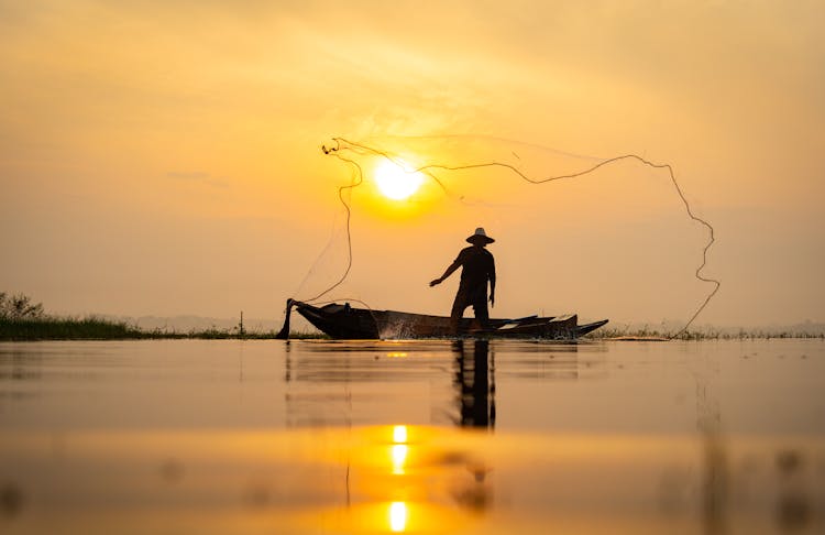 A Fisherman On A Boat Casting His Net At Sunrise