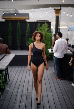 Elegant woman confidently walking in a stylish black swimsuit at an outdoor venue.