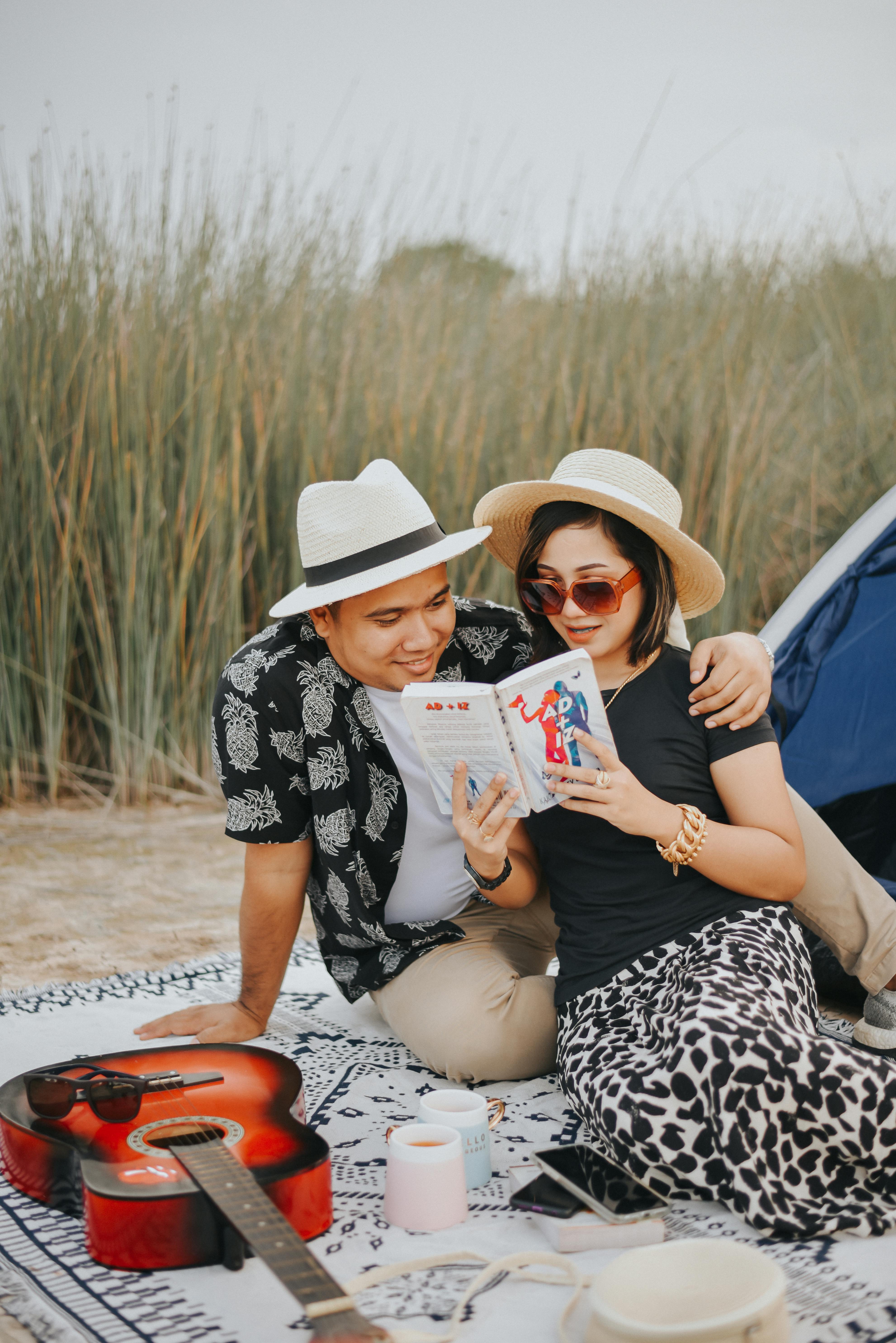 Couple enjoying a leisure reading time outdoors with a guitar at their side, wearing summer hats.