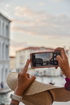 A person capturing the scenic Venice canal on a smartphone with a view of historic buildings during sunset.