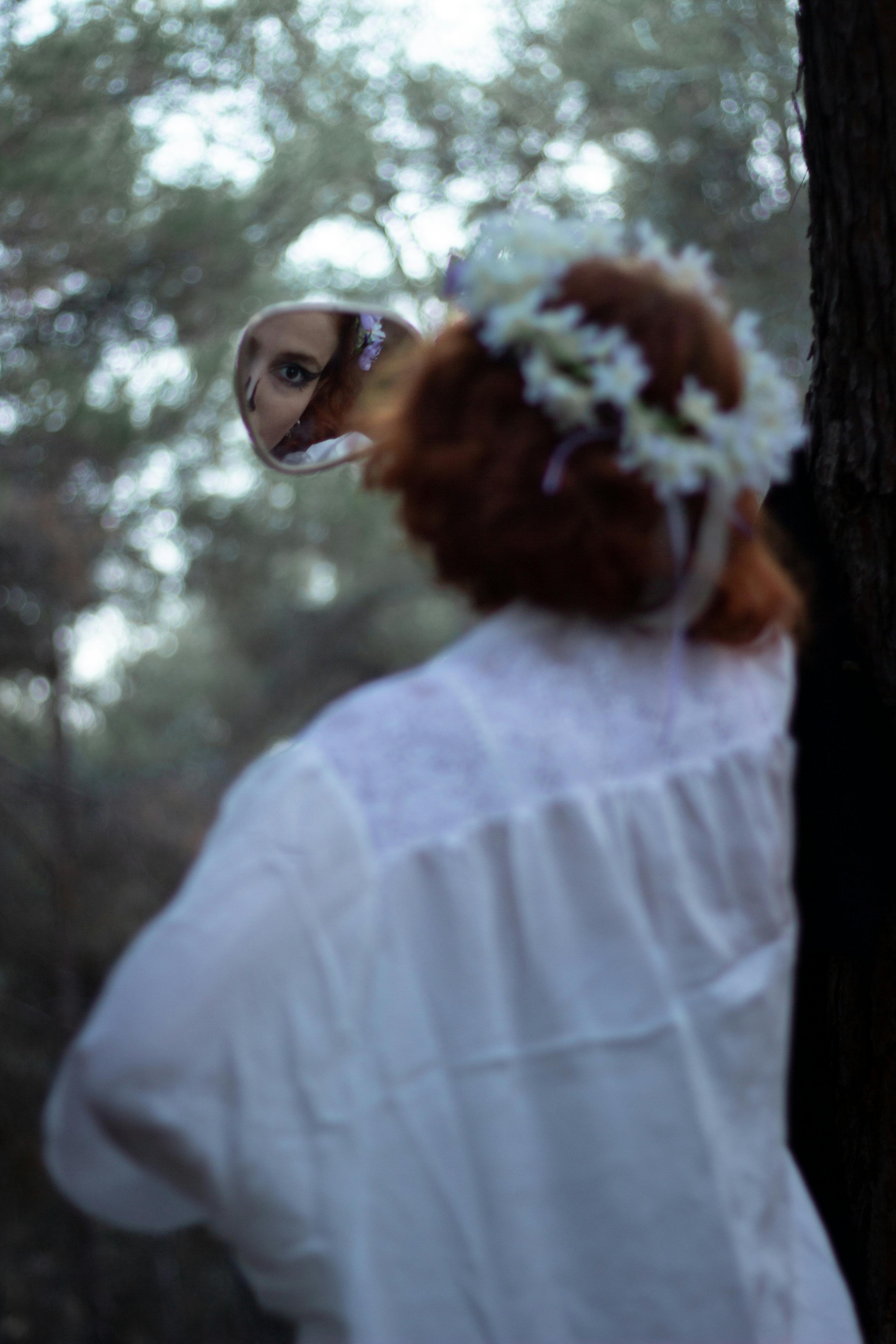 A woman's reflection in a handheld mirror, adorned with a floral wreath, in a serene forest setting.
