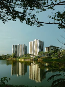 High-rise buildings reflecting on a serene lake under a clear blue sky, surrounded by lush greenery.