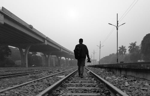 A Grayscale Photo of a Man Walking at the Railway