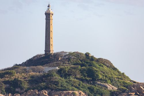 Free A towering lighthouse surrounded by lush greenery on a rocky hilltop under a clear blue sky. Stock Photo