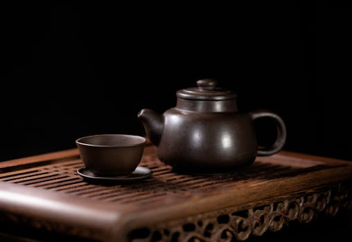 A minimalist still life of a ceramic teapot and cup on a wooden tray against a black background.