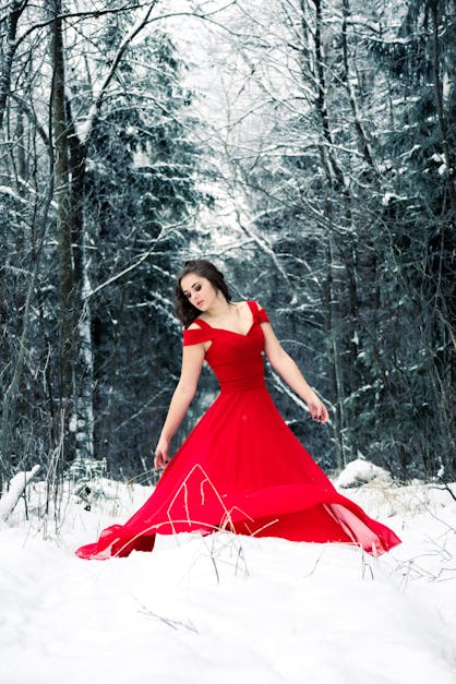 Young redhead girl in flowing red dress in forest with snowfall. stock photo