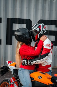 A couple in helmets and racing gear embraces on a motorcycle, symbolizing love and adventure.