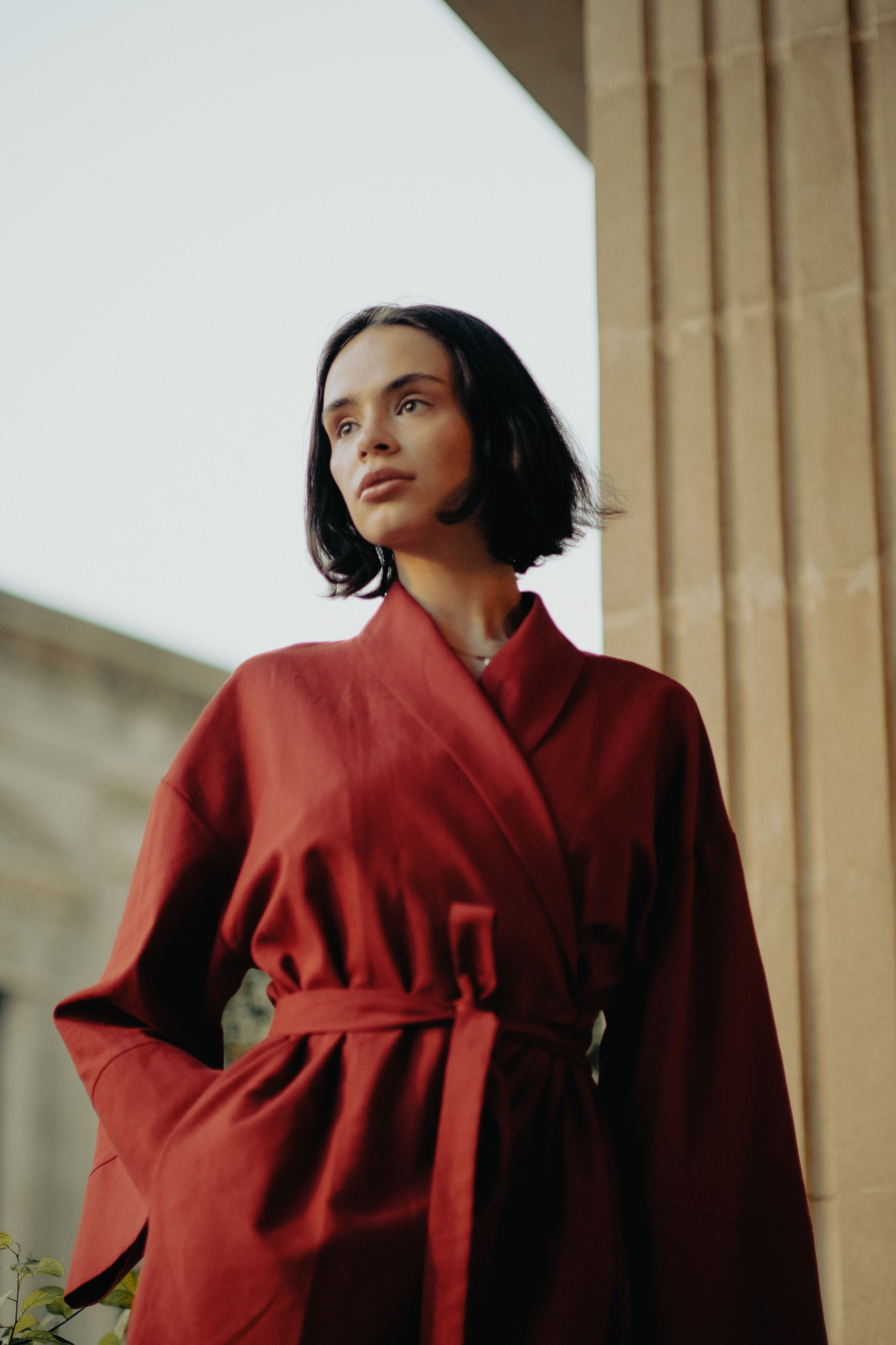 Mulher estilosa em um robe vermelho olhando pensativa para o exterior contra um fundo arquitetônico.