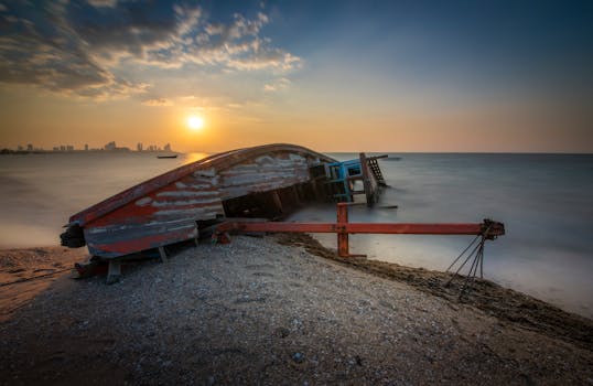 A derelict boat on a sandy beach with a vibrant sunset over the calm sea.