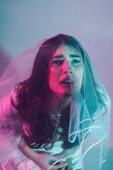 A distressed bride gazes upwards, draped in a veil and bathed in colorful lighting.