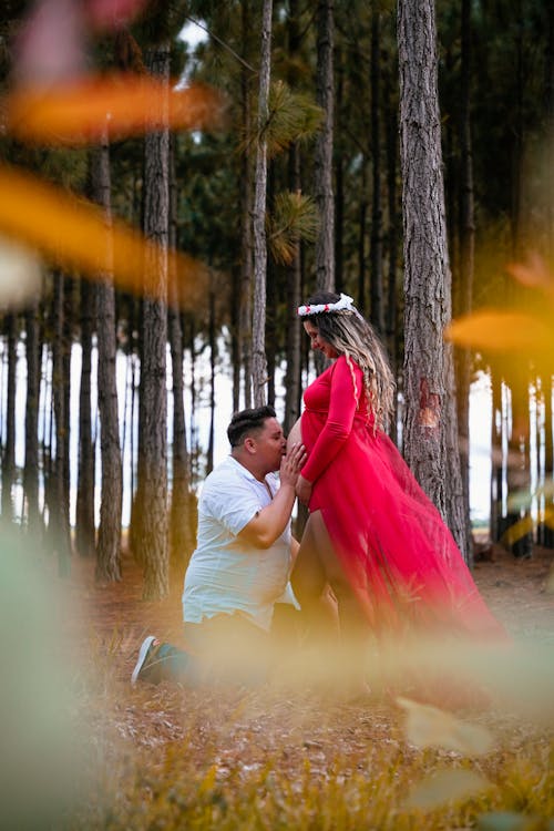 Man Kissing Pregnant Woman in Red Dress in Forest
