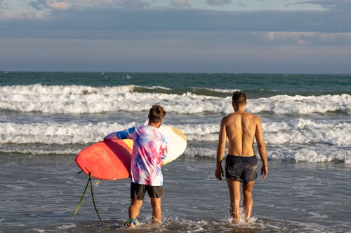 Back View of a Shirtless Man Walking beside a Man with a Surfboard