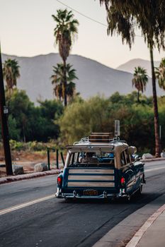 Classic vintage car driving on a palm-lined road against mountain backdrop at sunset.