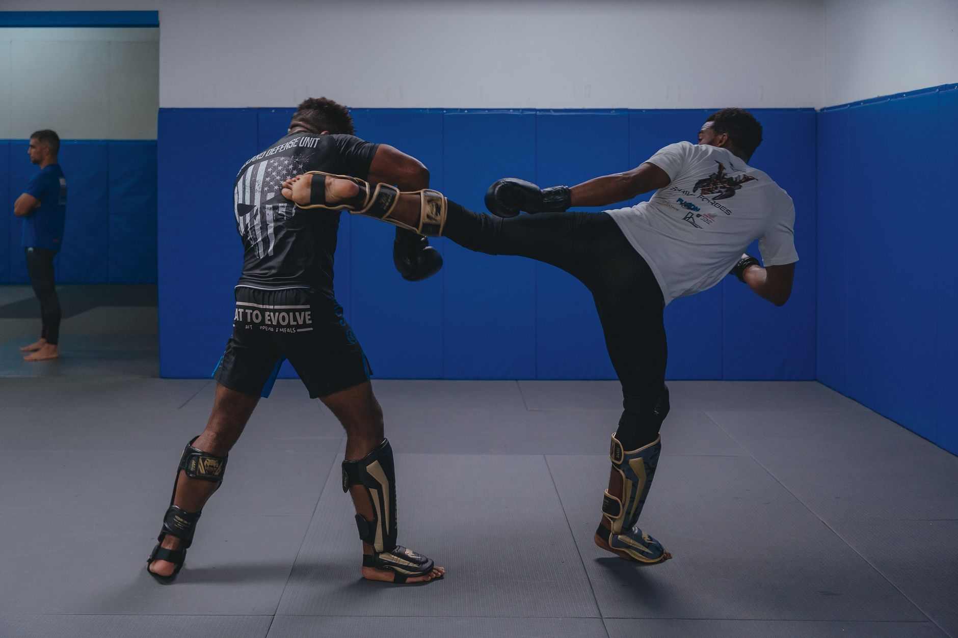 Two athletes practicing high kick techniques in an indoor martial arts gym environment.