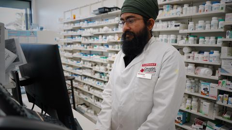 Pharmacist wearing a turban working at a computer in a pharmacy store with shelves filled with medicine.