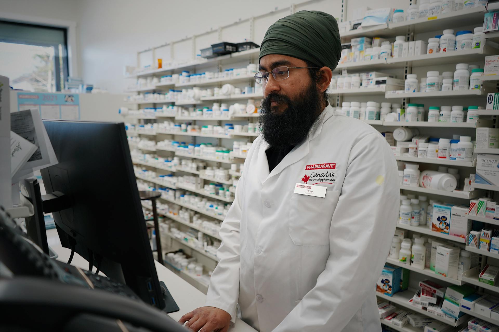 Pharmacist wearing a turban working at a computer in a pharmacy store with shelves filled with medicine.