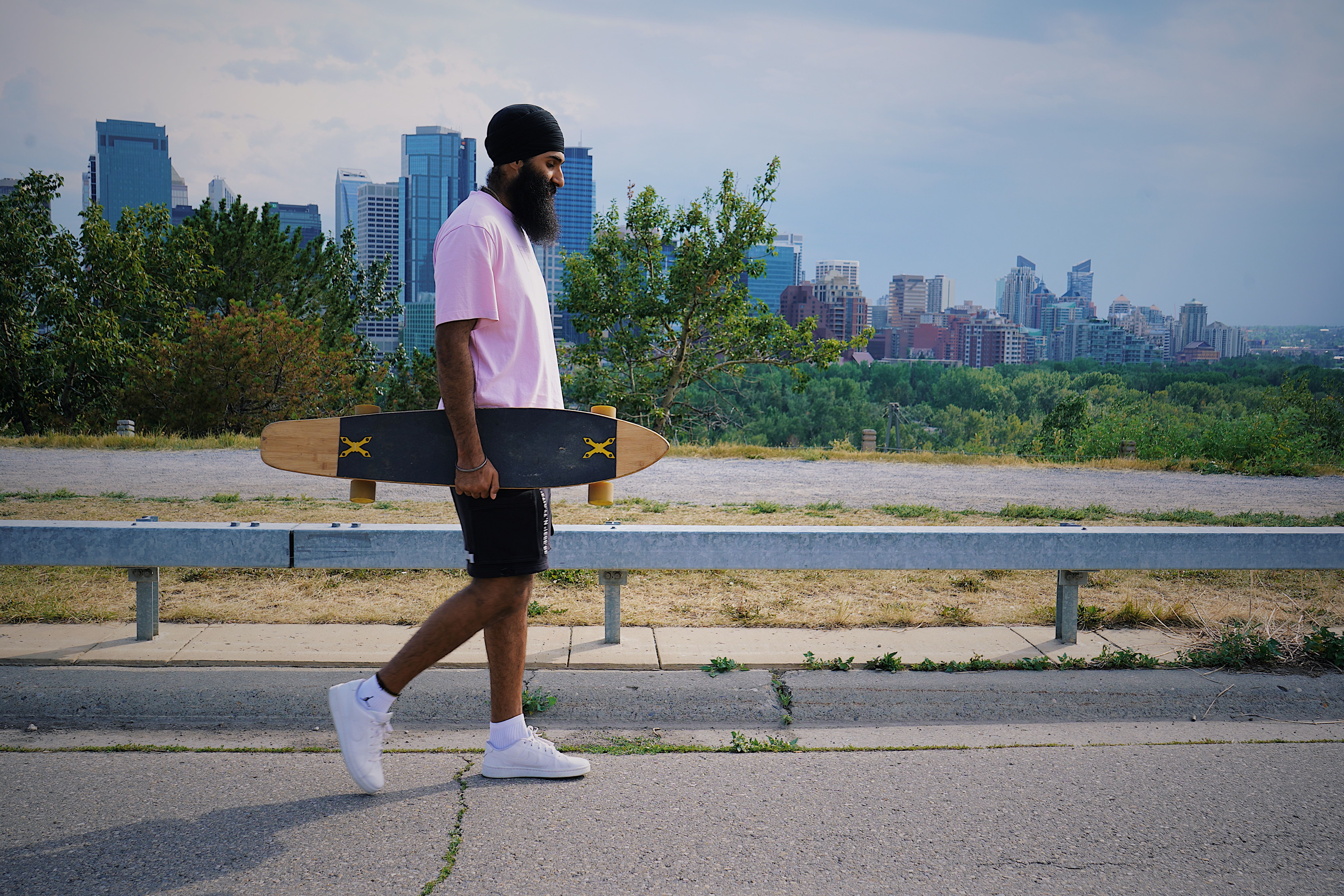 A man walking along a street carrying a longboard with a city skyline in the background.