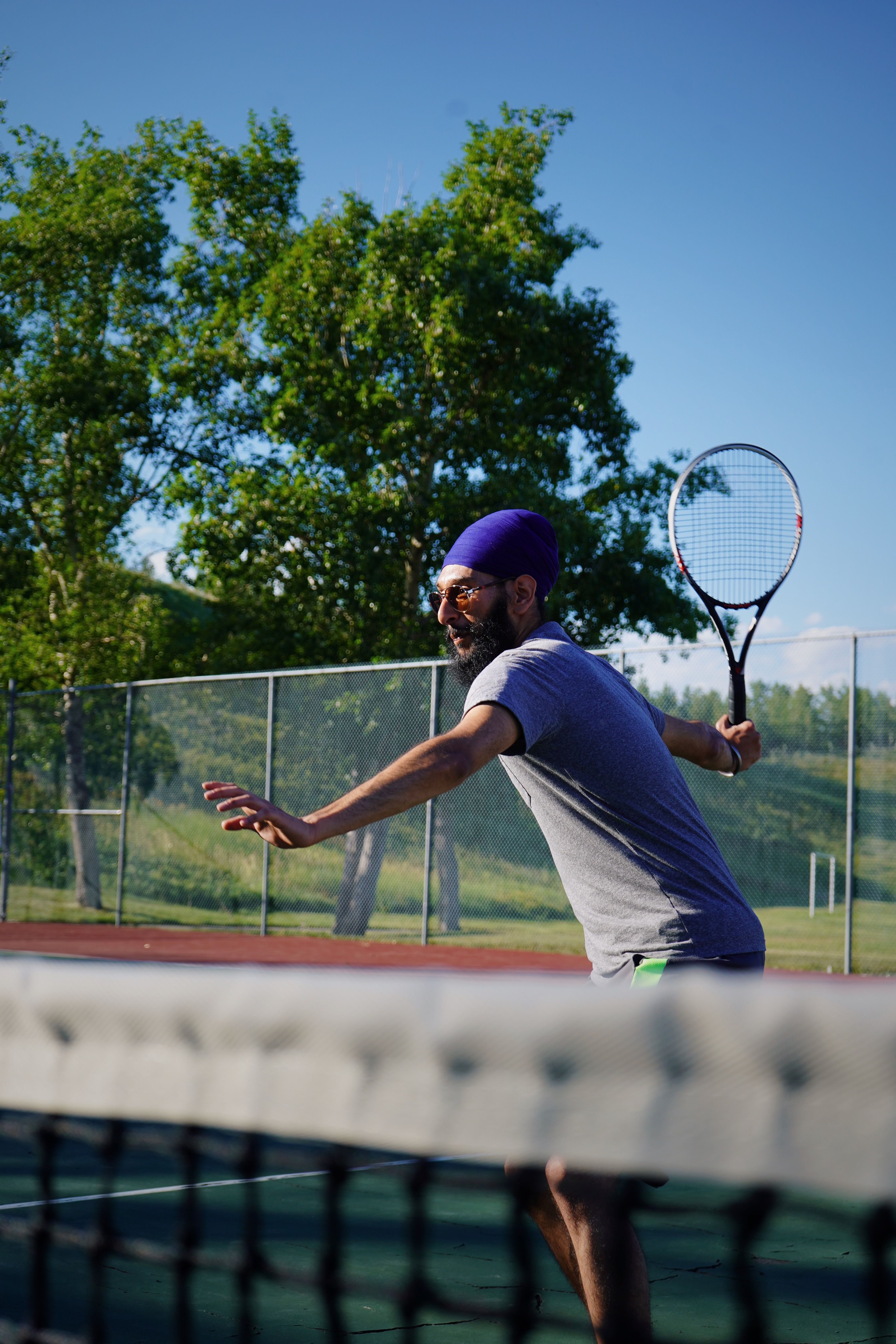 A man in mid-swing playing tennis on an outdoor court during a sunny summer day.