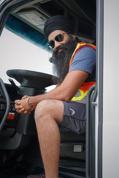 Truck driver with beard and sunglasses confidently poses in vehicle wearing safety vest.