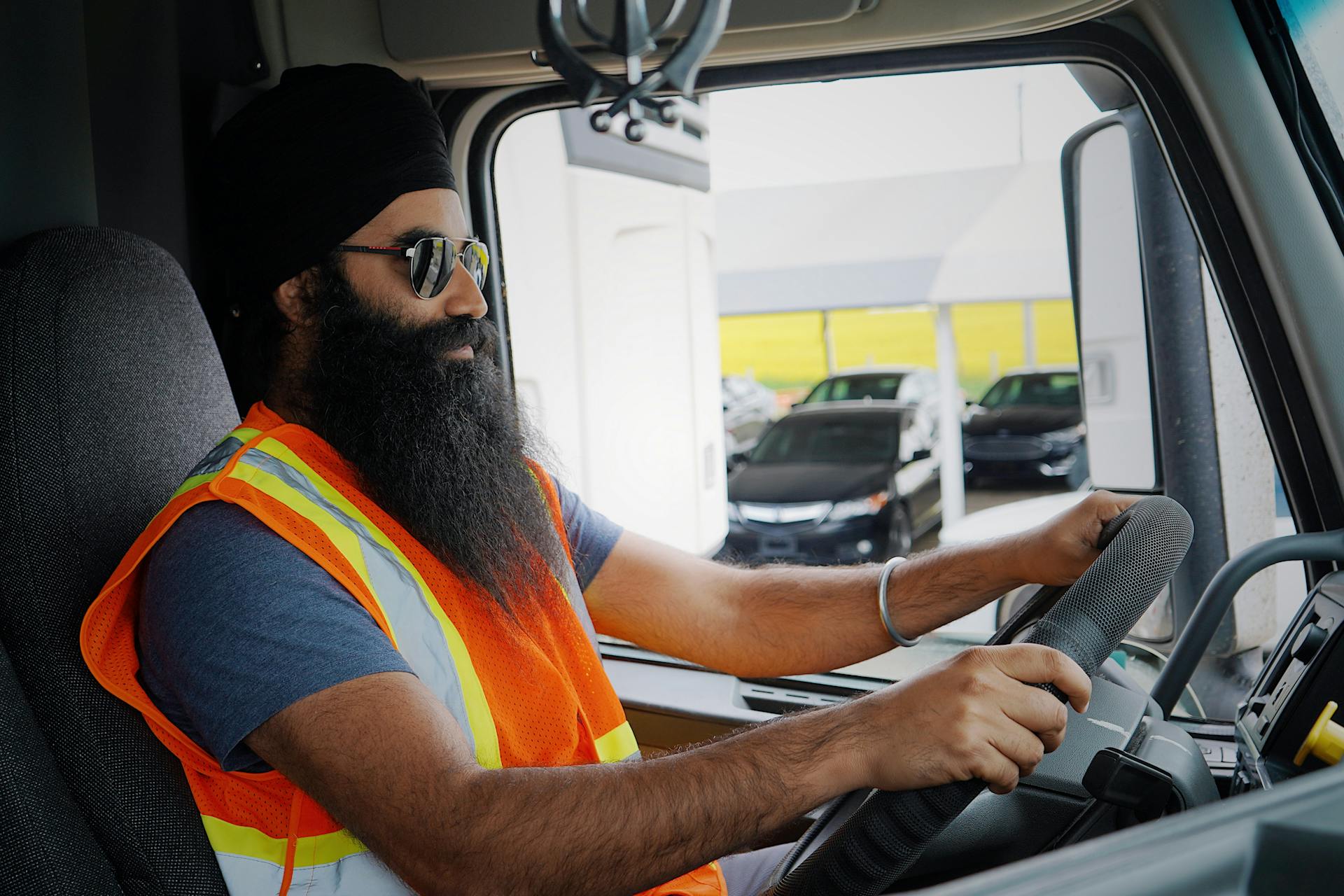 Bearded truck driver wearing a turban and safety vest inside a truck cab, focused on driving.