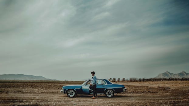 A man standing by a classic car in a barren landscape under a cloudy sky.