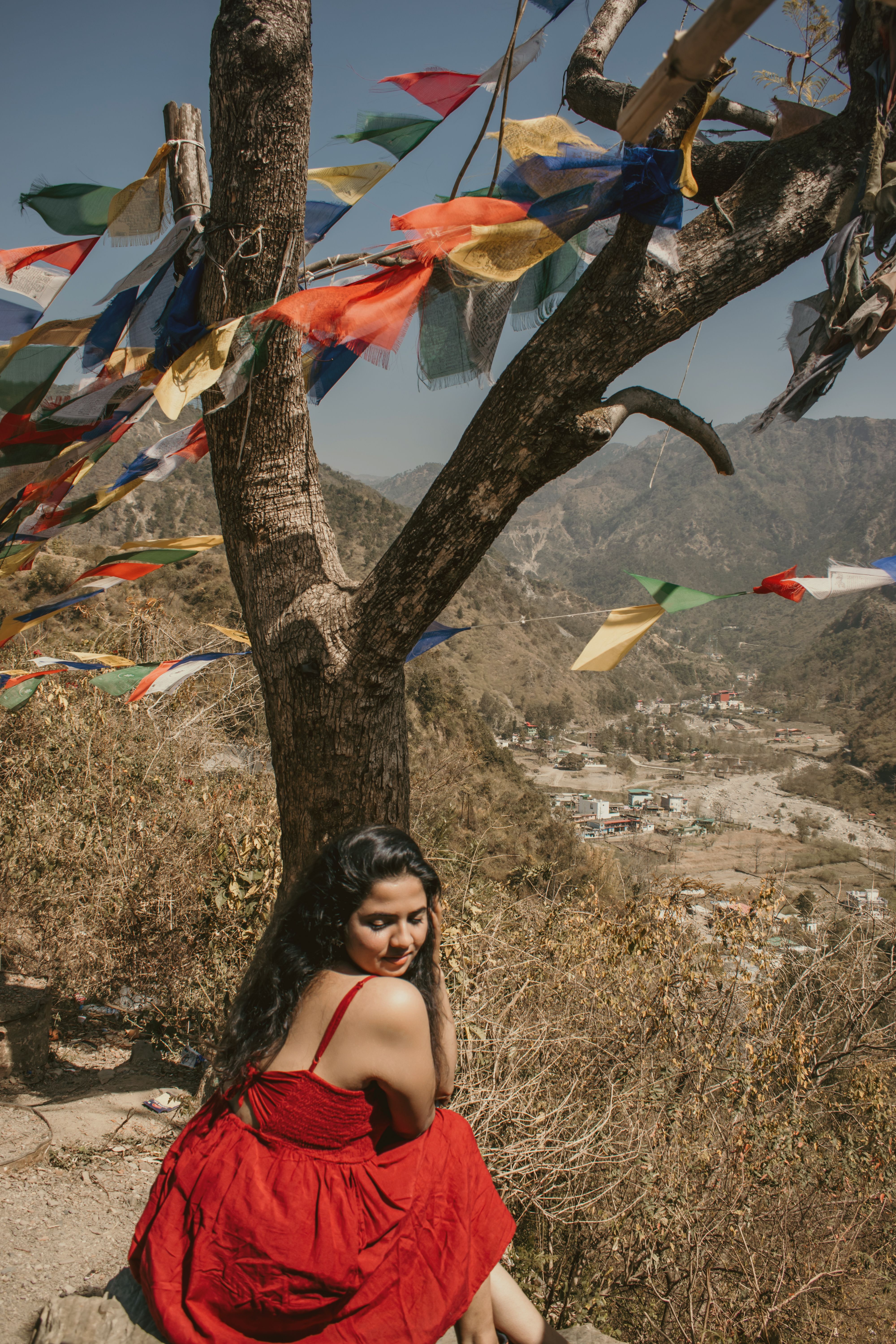 A young woman in a vibrant red dress sits by a tree adorned with prayer flags overlooking a scenic valley in Dehradun.