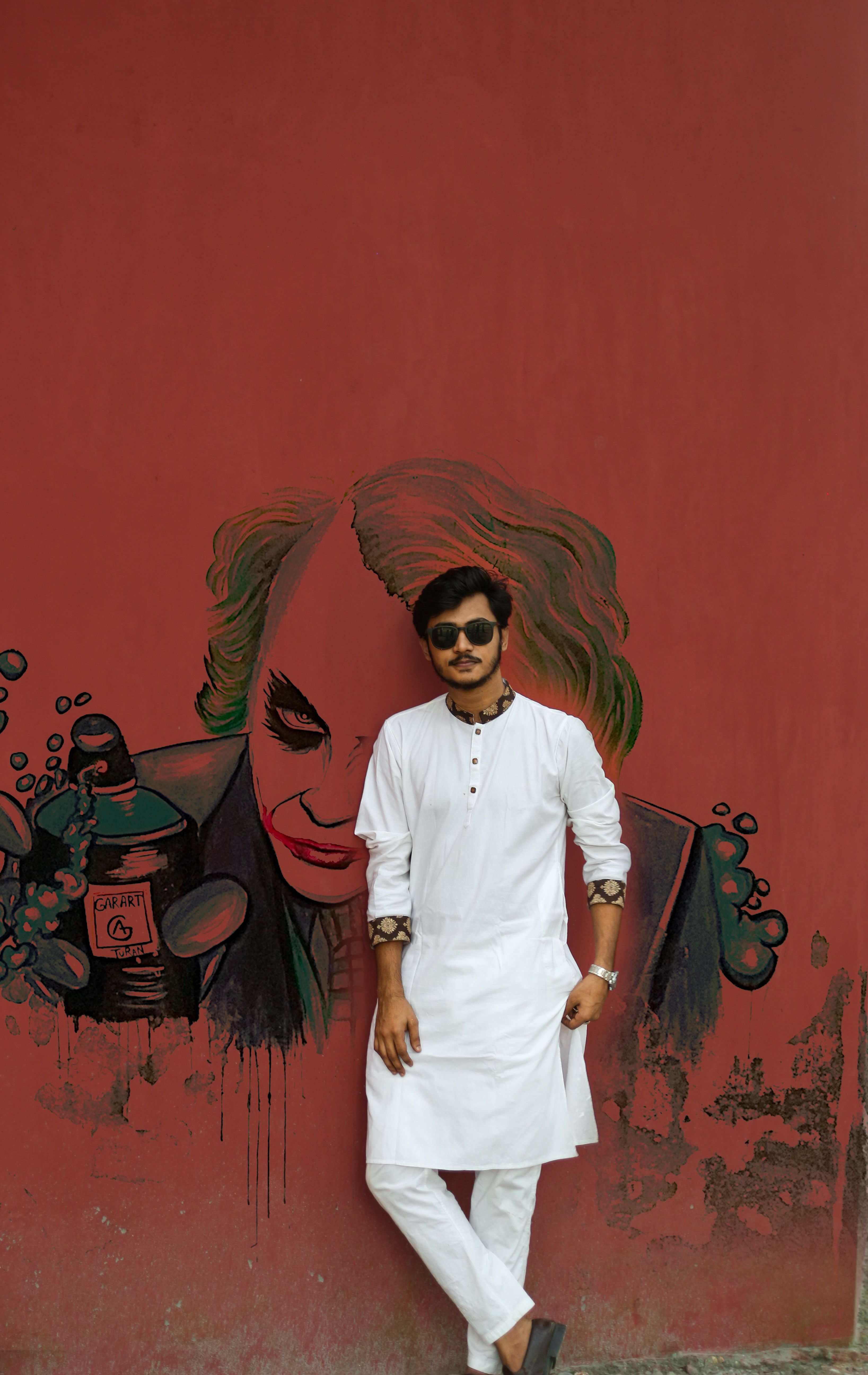 Man wearing a white sherwani leans against a graffiti-covered wall outdoors.
