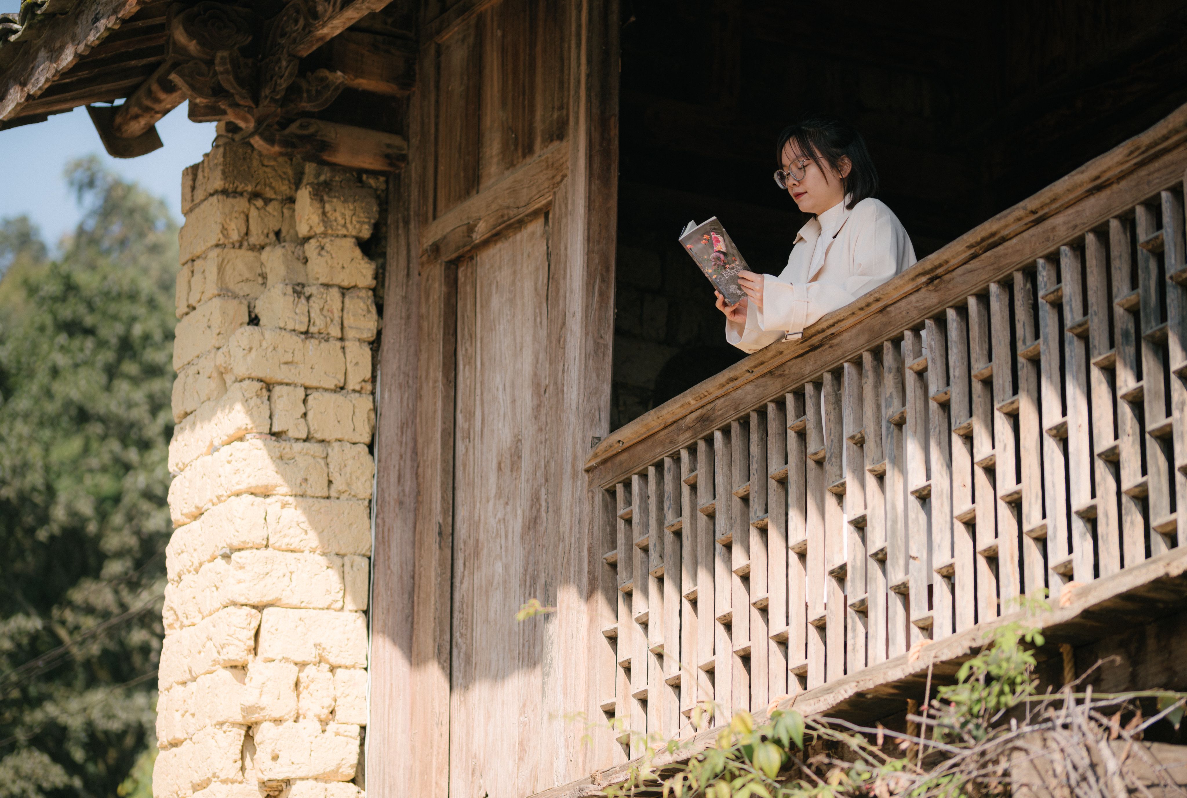 Woman wearing glasses reads a book on a rustic wooden balcony in sunlight.