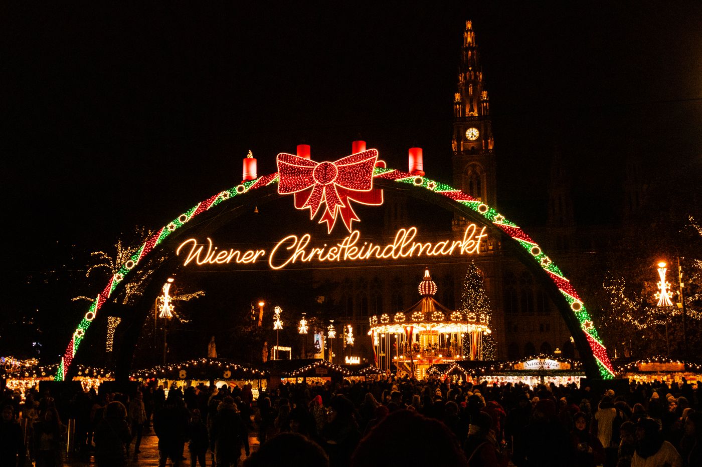 The illuminated Wiener Christkindlmarkt arch at night, with the market and Vienna City Hall tower behind
