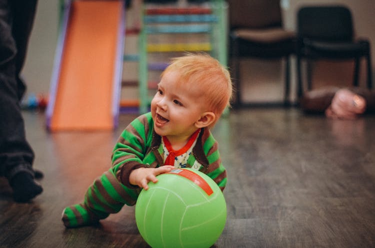 Cute Infant Holding A Green Ball On Wooden Floor