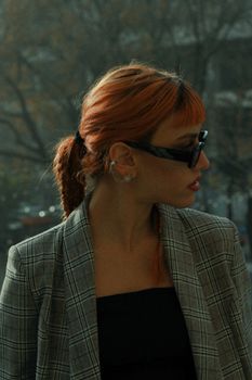 A fashionable woman with sunglasses and red hair looking sideways in a plaid blazer outdoors.