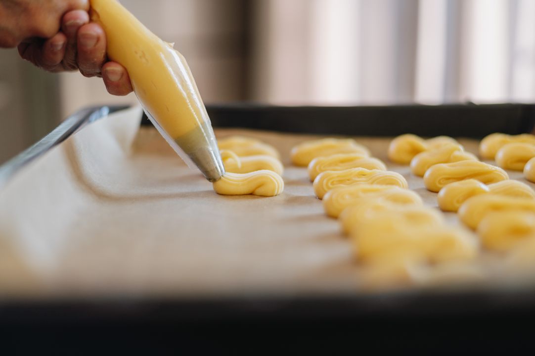 Choux Pastry Basics: Eclairs, Profiteroles, and Cream Puffs at Home 1 Choux pastry dough being piped from a pastry bag onto parchment-lined baking tray before baking