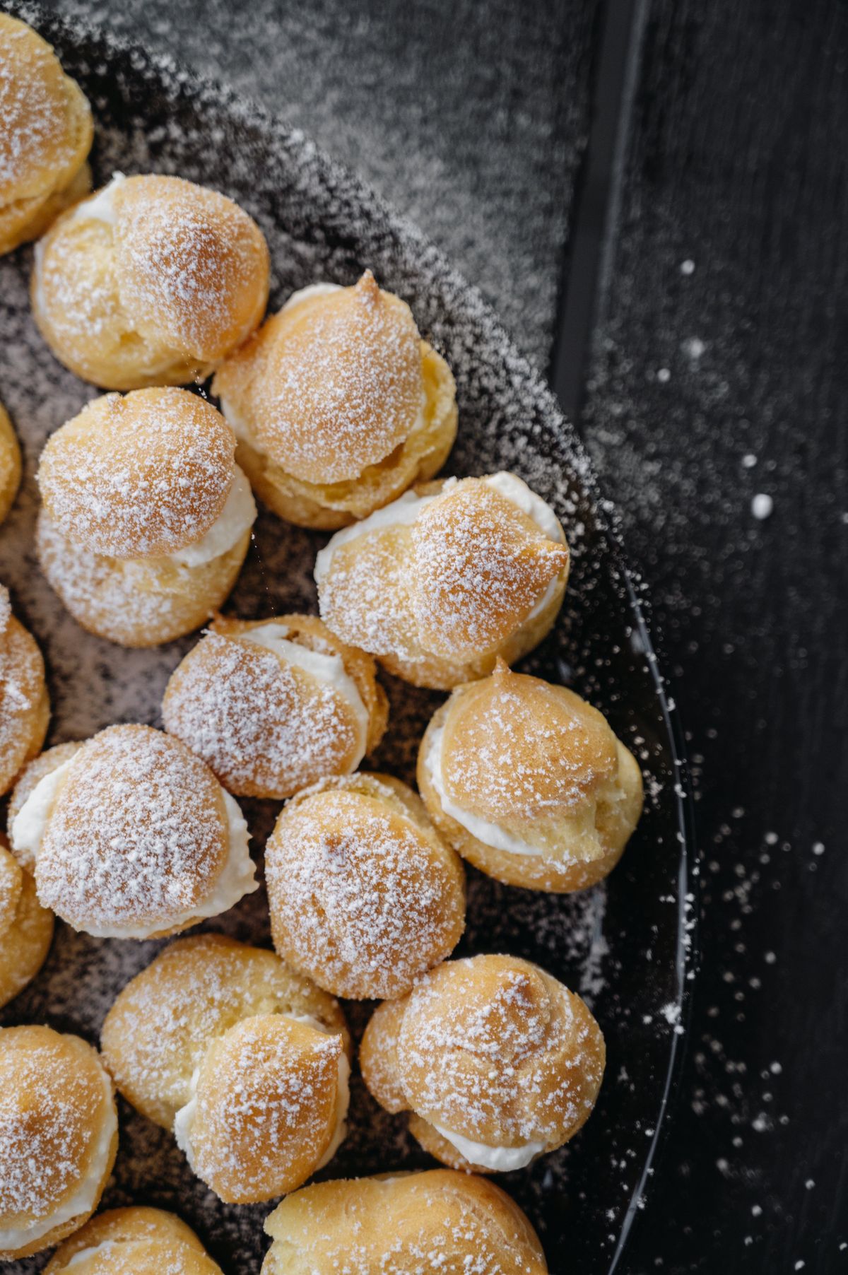 A heap of small sugared choux puffs on a dark tray.