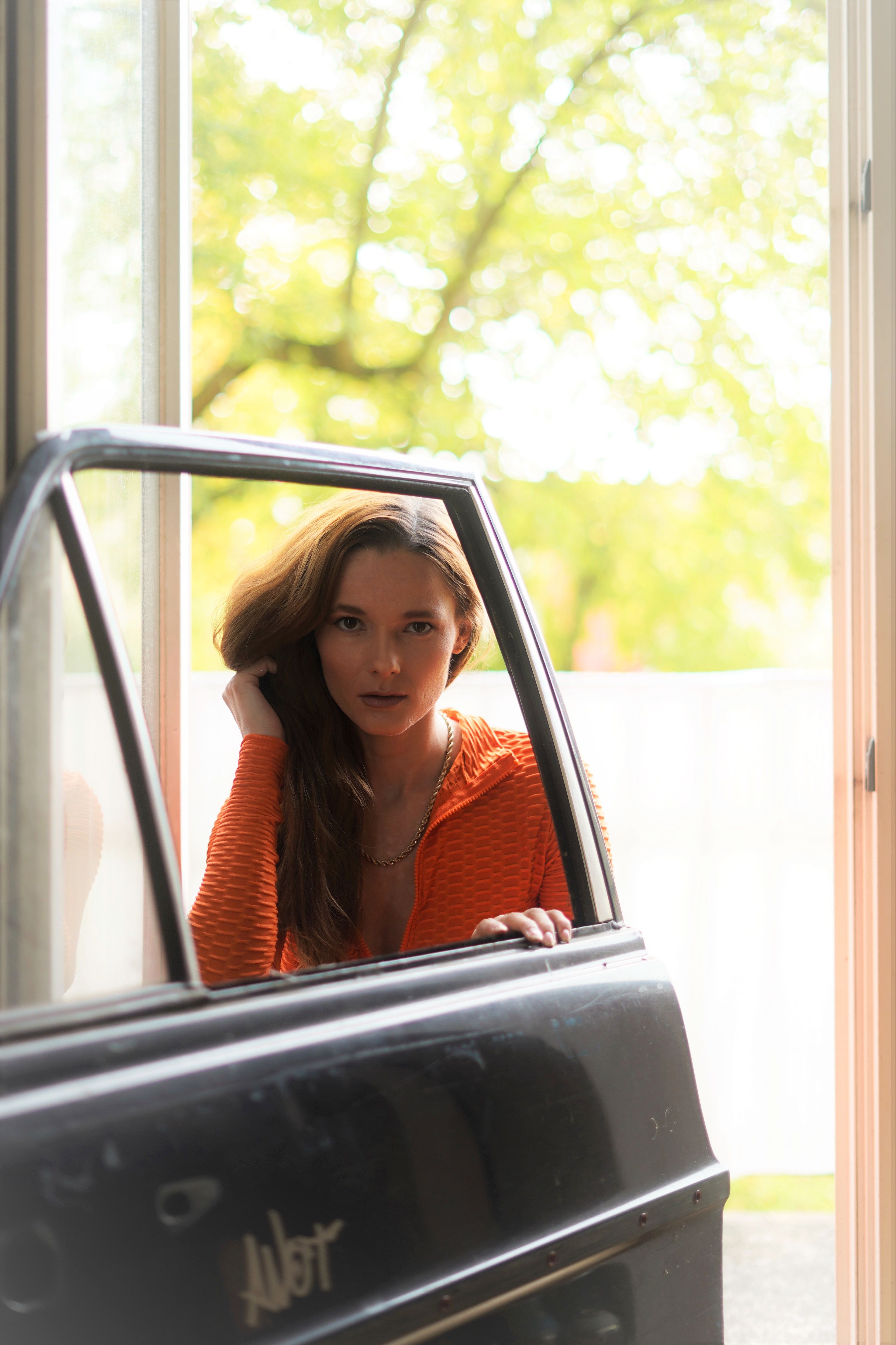 Elegant caucasian woman in orange jacket posing at a vintage car door with a blurred summer background.