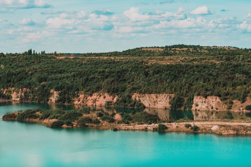 Free A tranquil view of a blue lake surrounded by lush forest and hills under a bright sky. Stock Photo