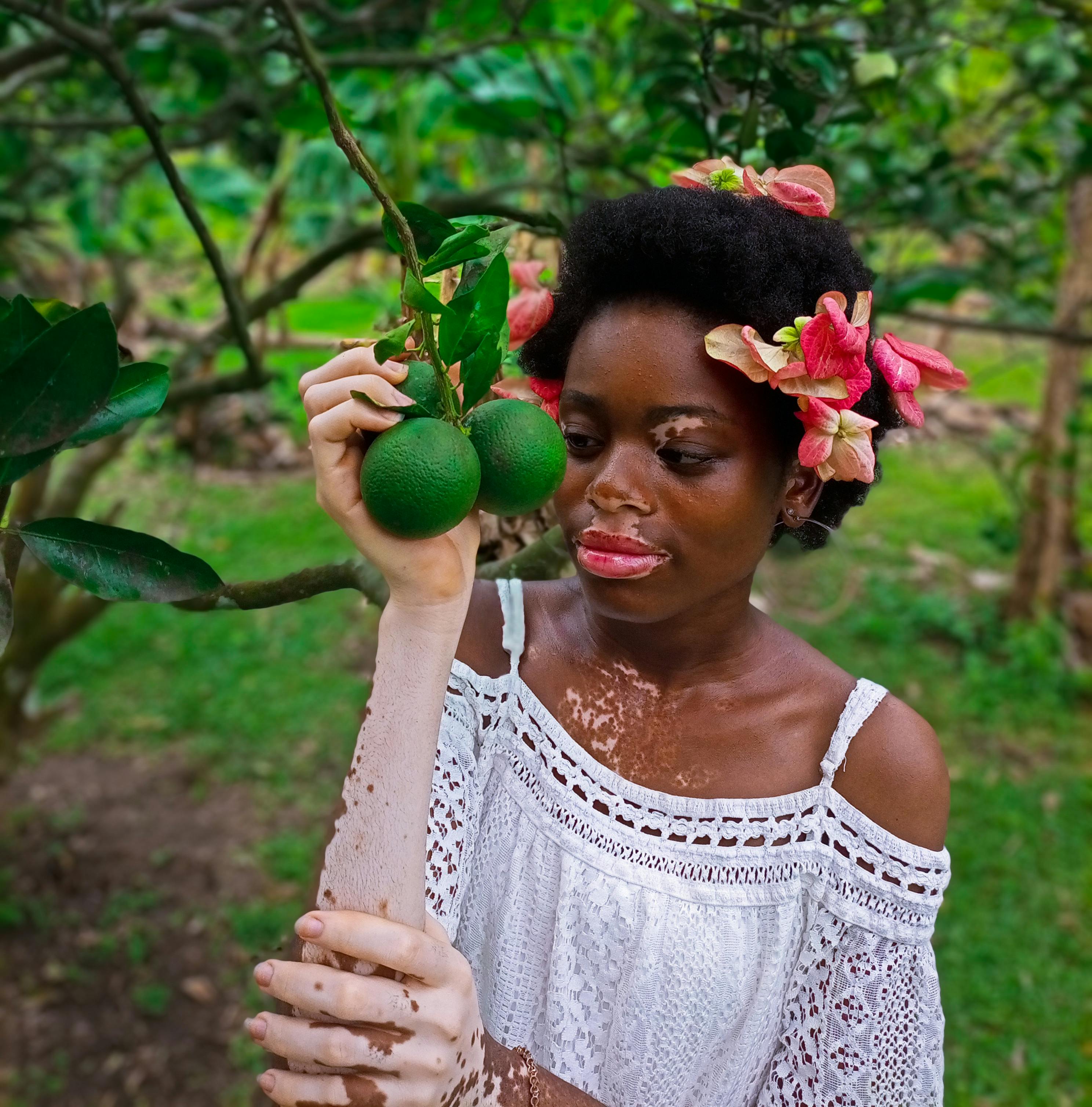 Portrait of a woman with floral hair accessories holding green oranges in a lush garden.