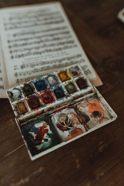 A creative composition of watercolor paints and music notes, featuring engagement rings on a wooden table.