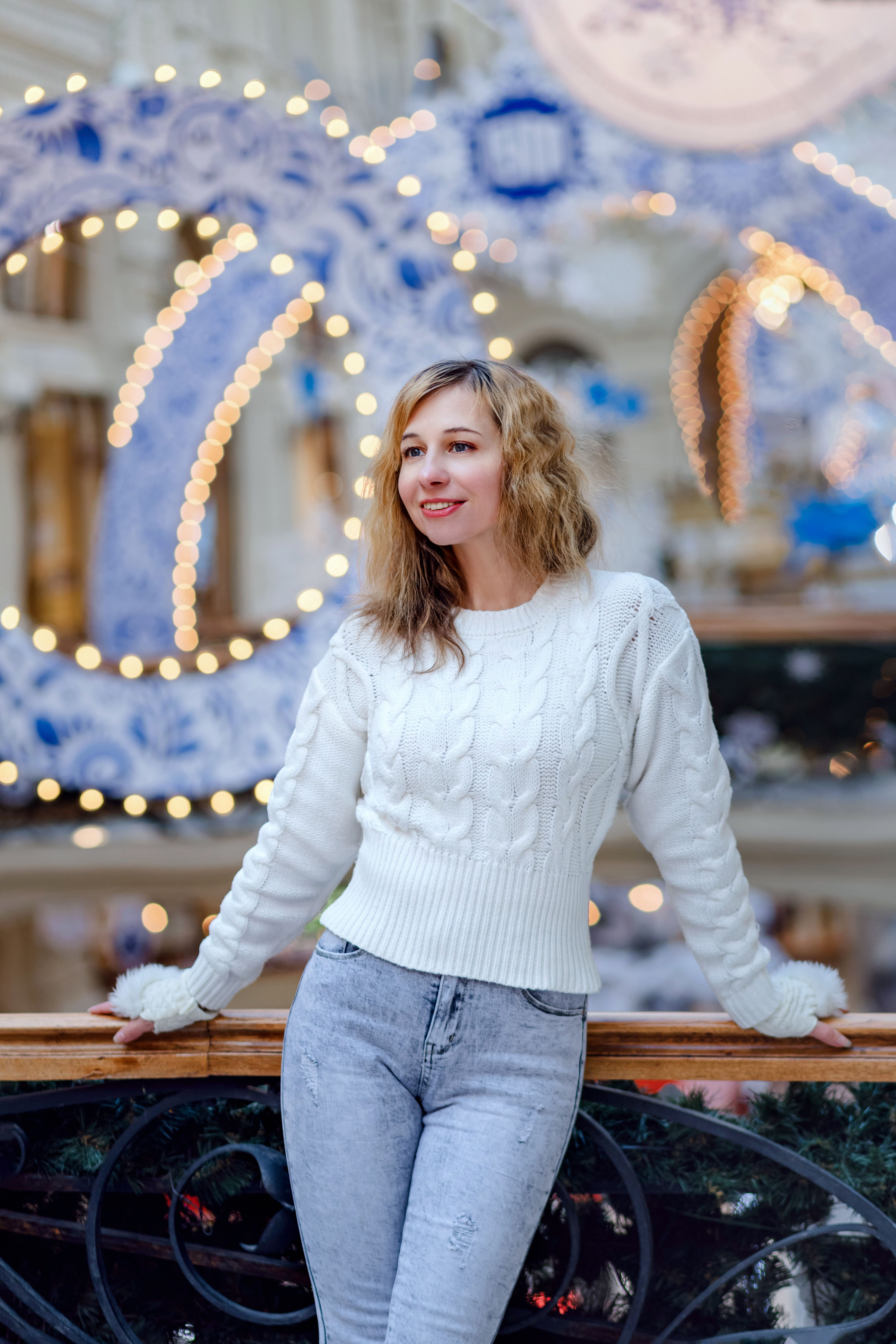 Caucasian woman in a white sweater smiling at an indoor festive setting with lights.