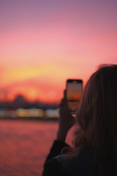 A woman takes a photo of a dramatic sunset with her smartphone, silhouetted against a vibrant pink sky.