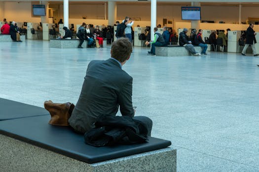 Businessman in suit seated in airport terminal waiting area, observing travelers.